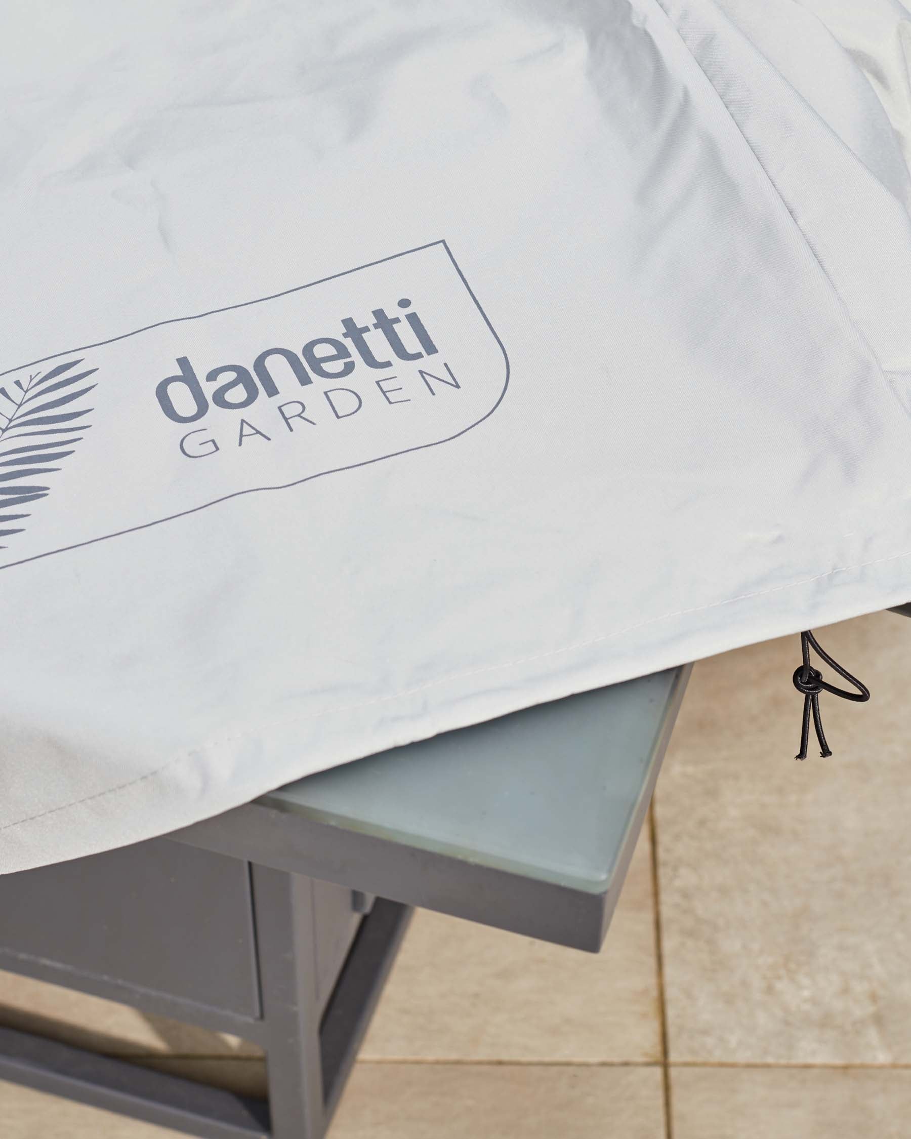 A close-up of a modern outdoor cushion with white fabric, featuring the "Danetti Garden" logo, placed on a frosted glass tabletop supported by a sleek grey metal frame.