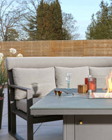 Modern outdoor furniture set featuring a sleek, grey metal frame sofa with light grey cushions and a matching grey metal coffee table with a smooth top. Accessories include a clear drinking glass, sunglasses, and decorative candles, positioned on the coffee table which is next to the sofa. The furniture is set against a backdrop of a wooden privacy fence and greenery.