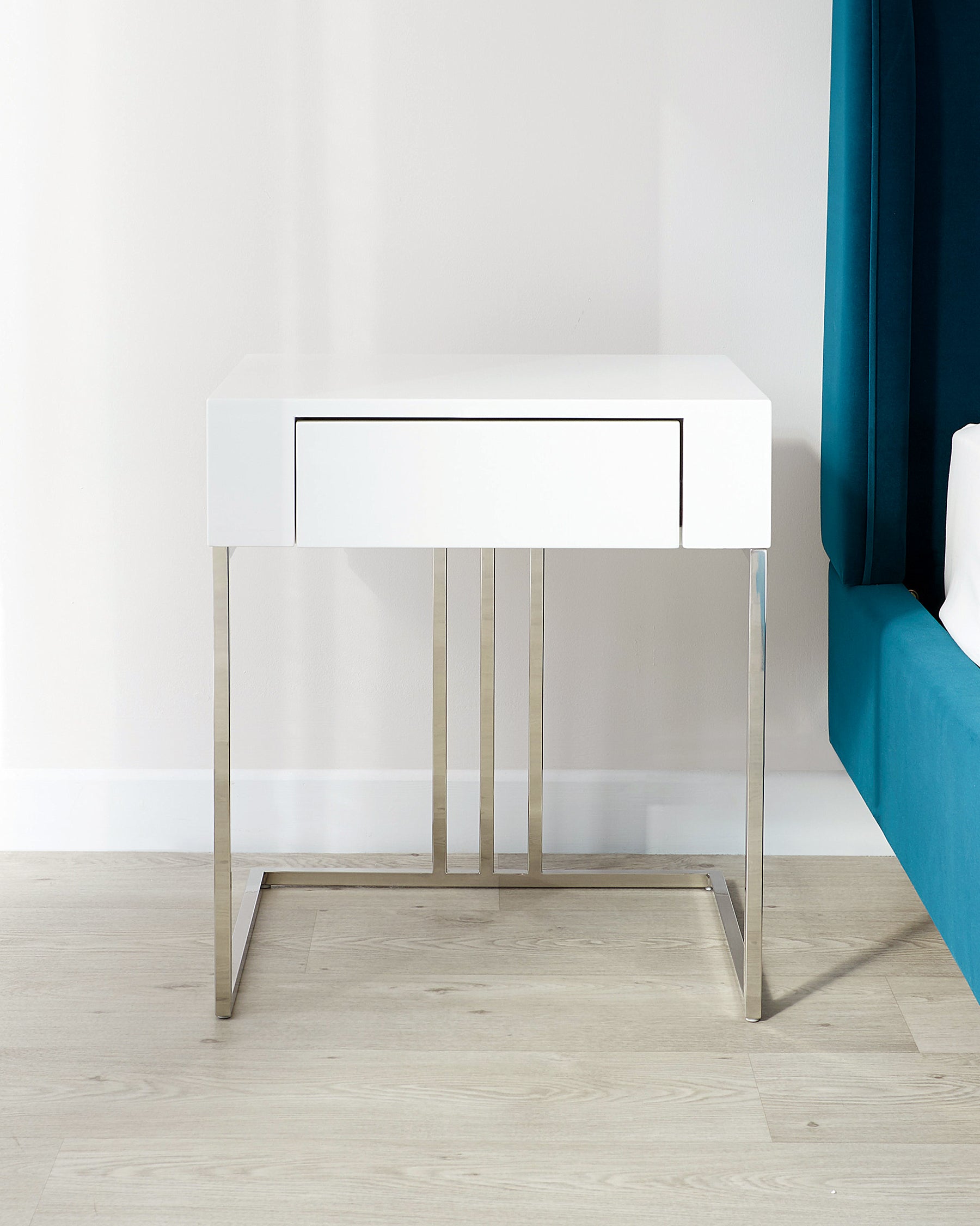 A modern white lacquer console table with a sleek drawer and polished stainless steel legs, positioned against a neutral wall beside a teal blue upholstered bed or sofa.