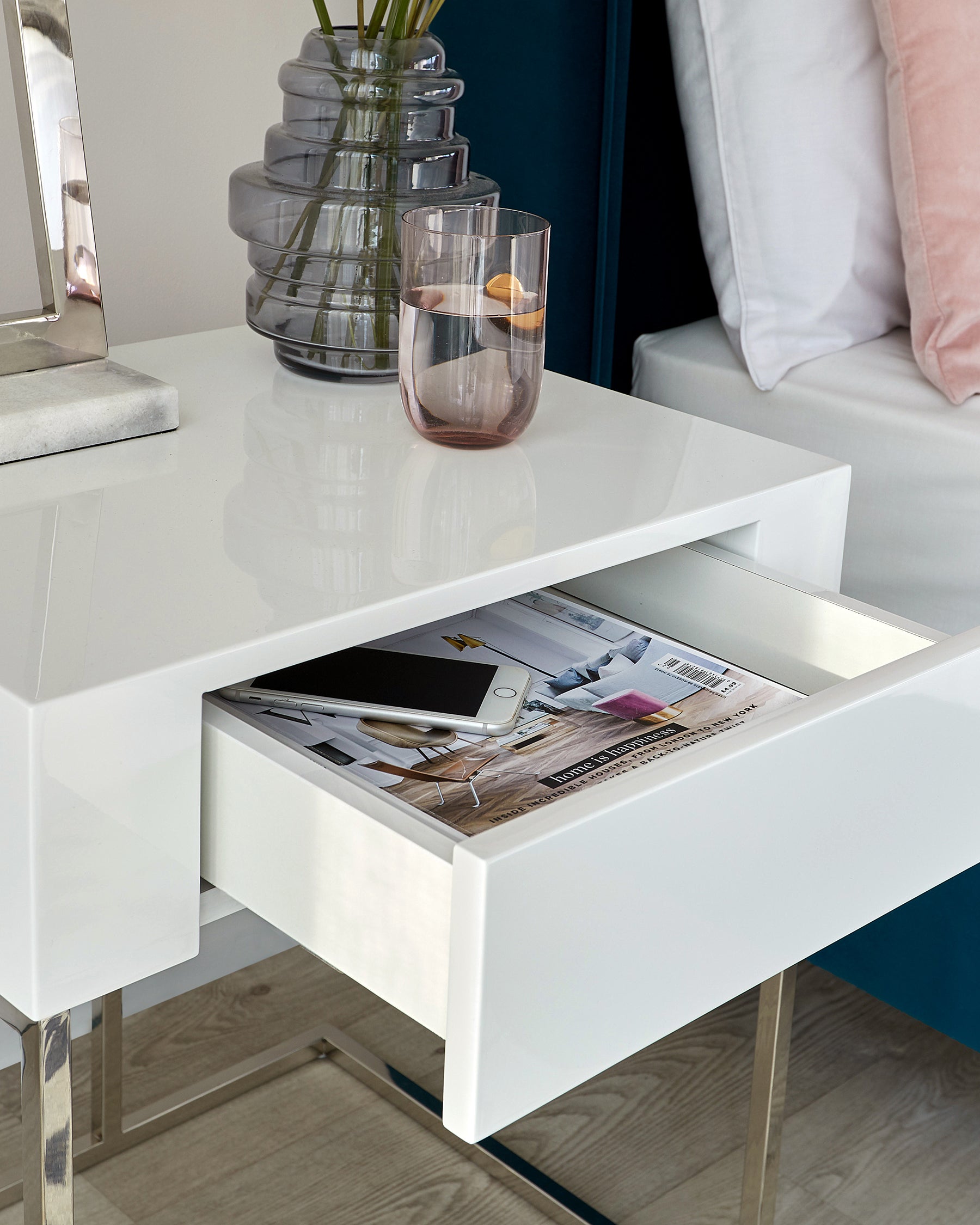 Glossy white modern side table with an open drawer filled with magazines, beside a sofa with a pale pink cushion, and a clear vase and glass on top of the table.