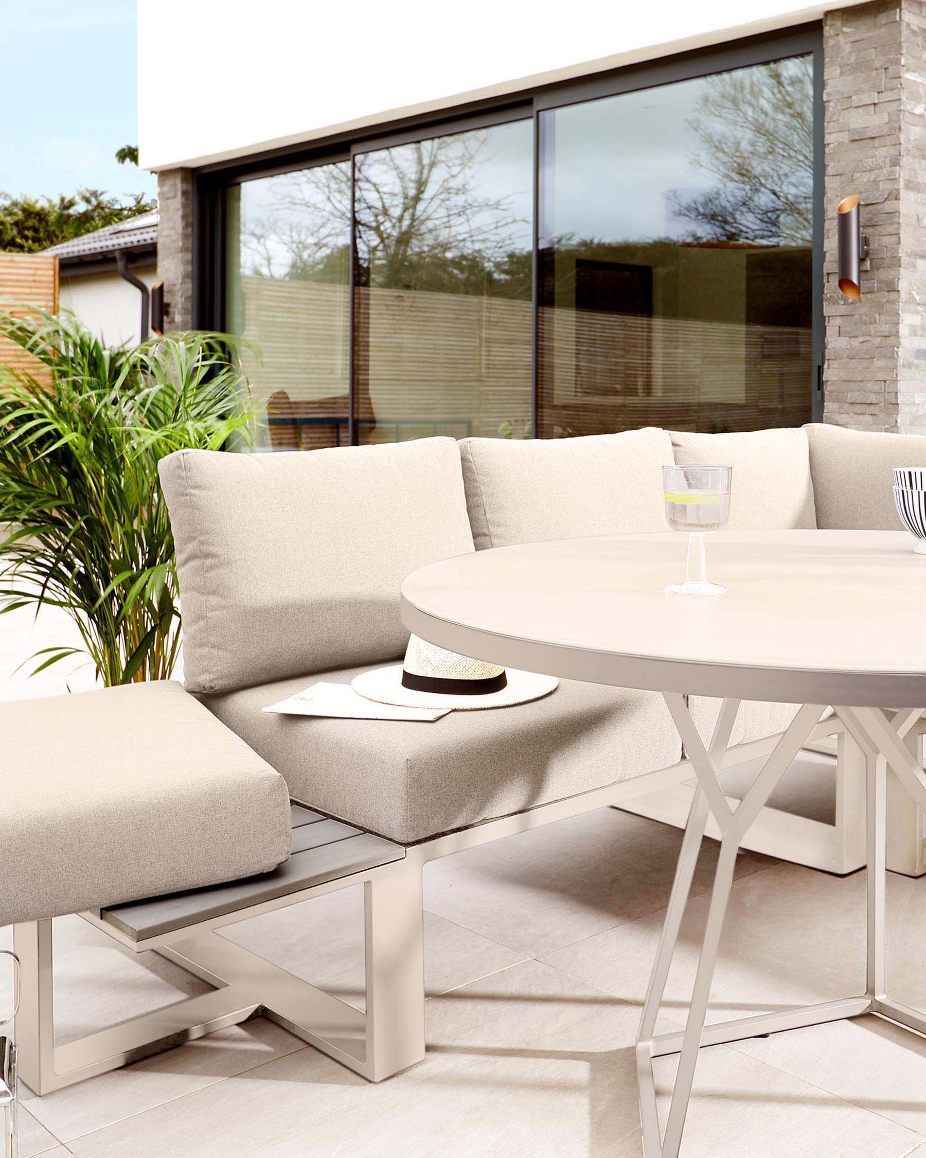 Modern outdoor furniture set featuring a neutral-toned sofa with plush cushions, a matching ottoman, and a round, white metal table with a stylish X-shaped base.