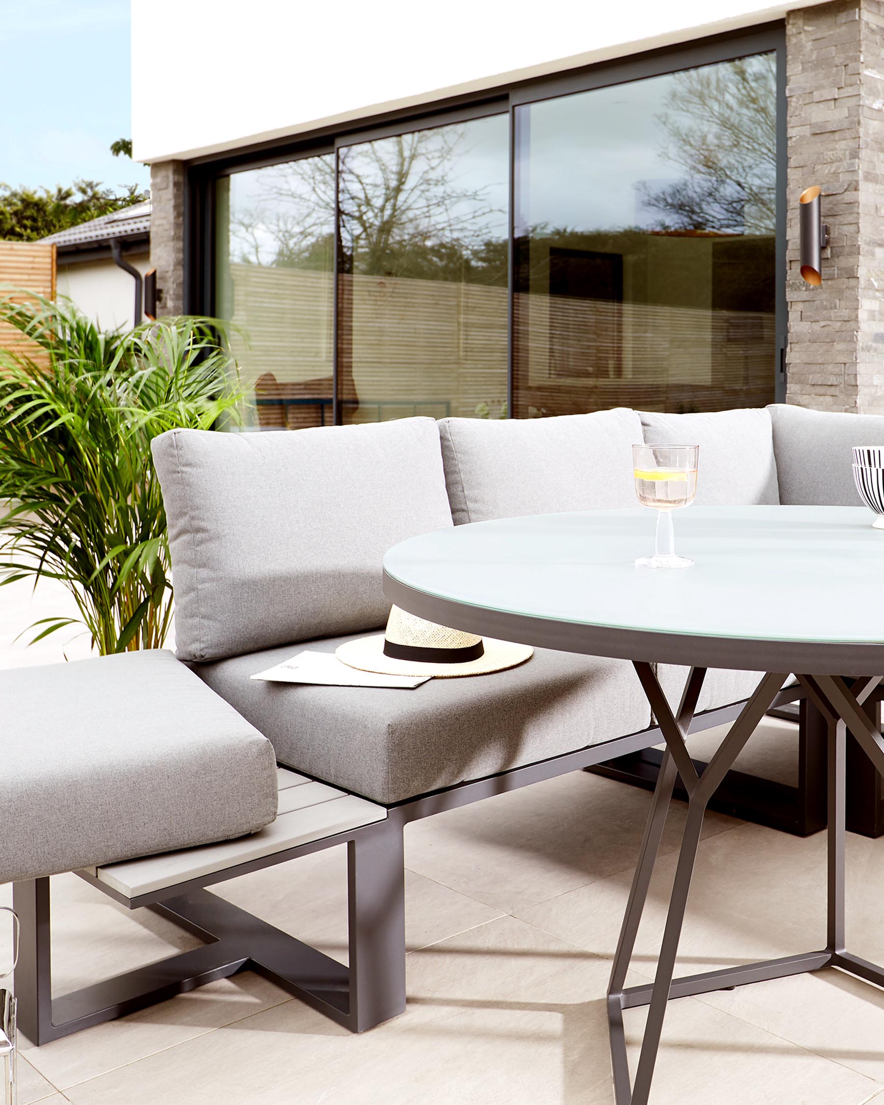 Modern outdoor furniture set featuring a sectional corner sofa with light grey cushions, a matching ottoman, and a round, white-top dining table with a sleek, dark metallic frame.