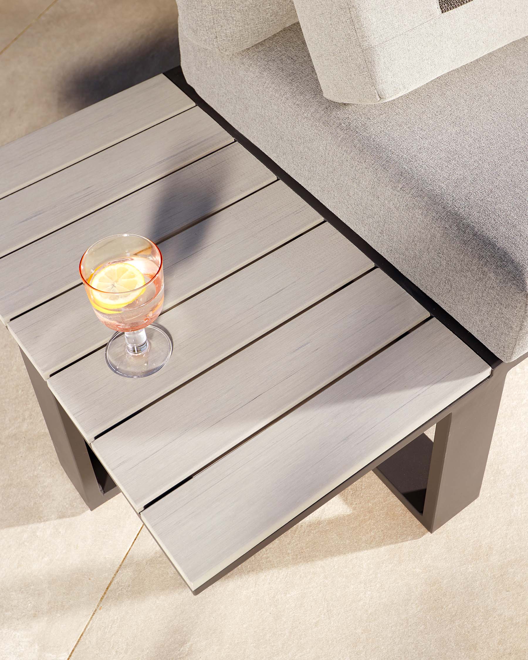 Contemporary outdoor side table with a rectangular top comprising wide, grey slatted boards and sturdy, A-frame legs, positioned next to a section of a grey upholstered sofa with a glass of beverage on its surface.