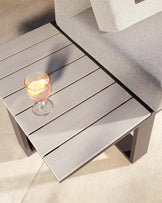 Modern outdoor grey slatted coffee table with a lower shelf, constructed from weather-resistant materials. The edge of a matching grey fabric sofa is visible, suggesting a coordinated set. A clear glass with a beverage and lemon slice rests on the table, highlighting its use in a patio or leisure setting.