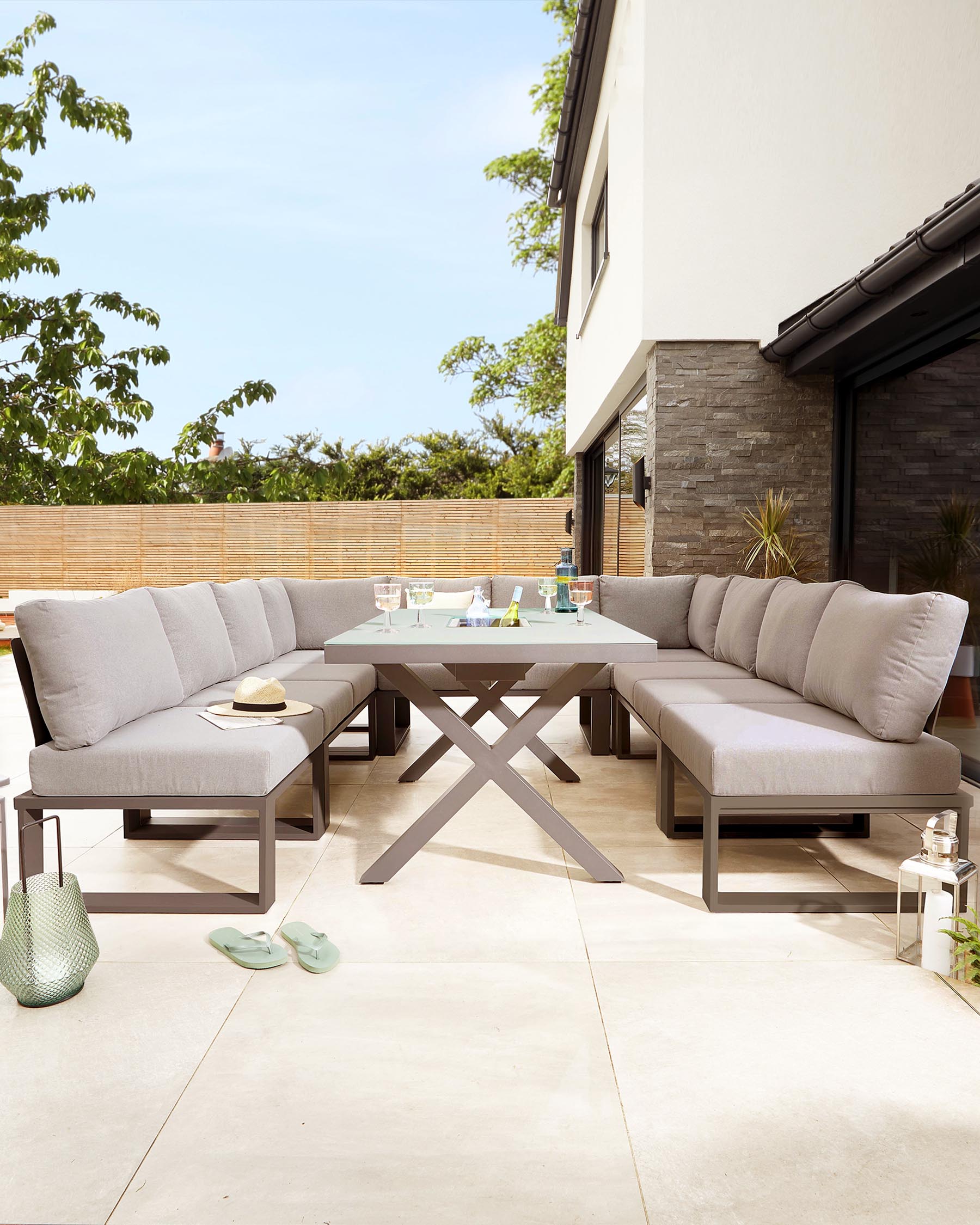 Contemporary outdoor patio furniture featuring a modular corner sofa with grey cushions and a rectangular dining table with an X-shaped base. The furniture frames are grey metal, harmonizing with neutral-toned cushions to create an elegant and comfortable outdoor dining and lounging area.