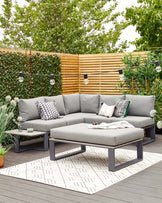 Modern outdoor patio furniture set featuring a sectional sofa with light grey cushions on a sleek, dark-framed base, complemented by matching grey throw pillows with various patterns. A rectangular ottoman serves as a central coffee table, mirroring the sofa's design. Flanking the sofa is a small square side table in a matching dark frame with a grey top. The set is arranged on a geometric-patterned outdoor rug, creating a cosy and stylish entertainment area.