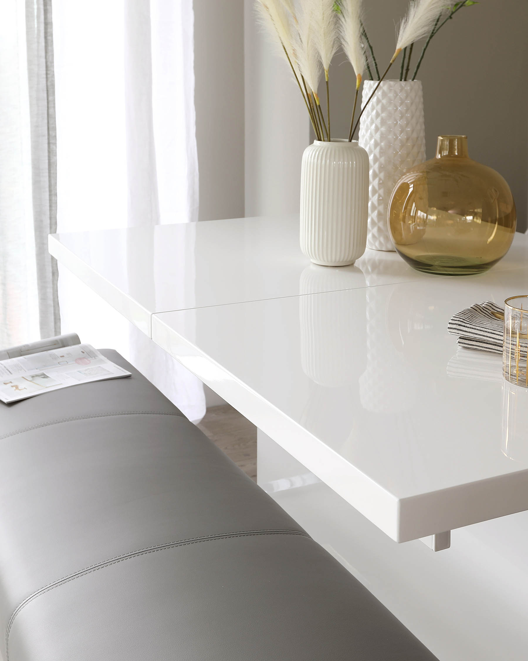 Modern minimalist design with a sleek white glossy rectangular dining table featuring clean lines and simple leg supports. A soft grey upholstered bench aligns with the table, complementing the contemporary aesthetic.