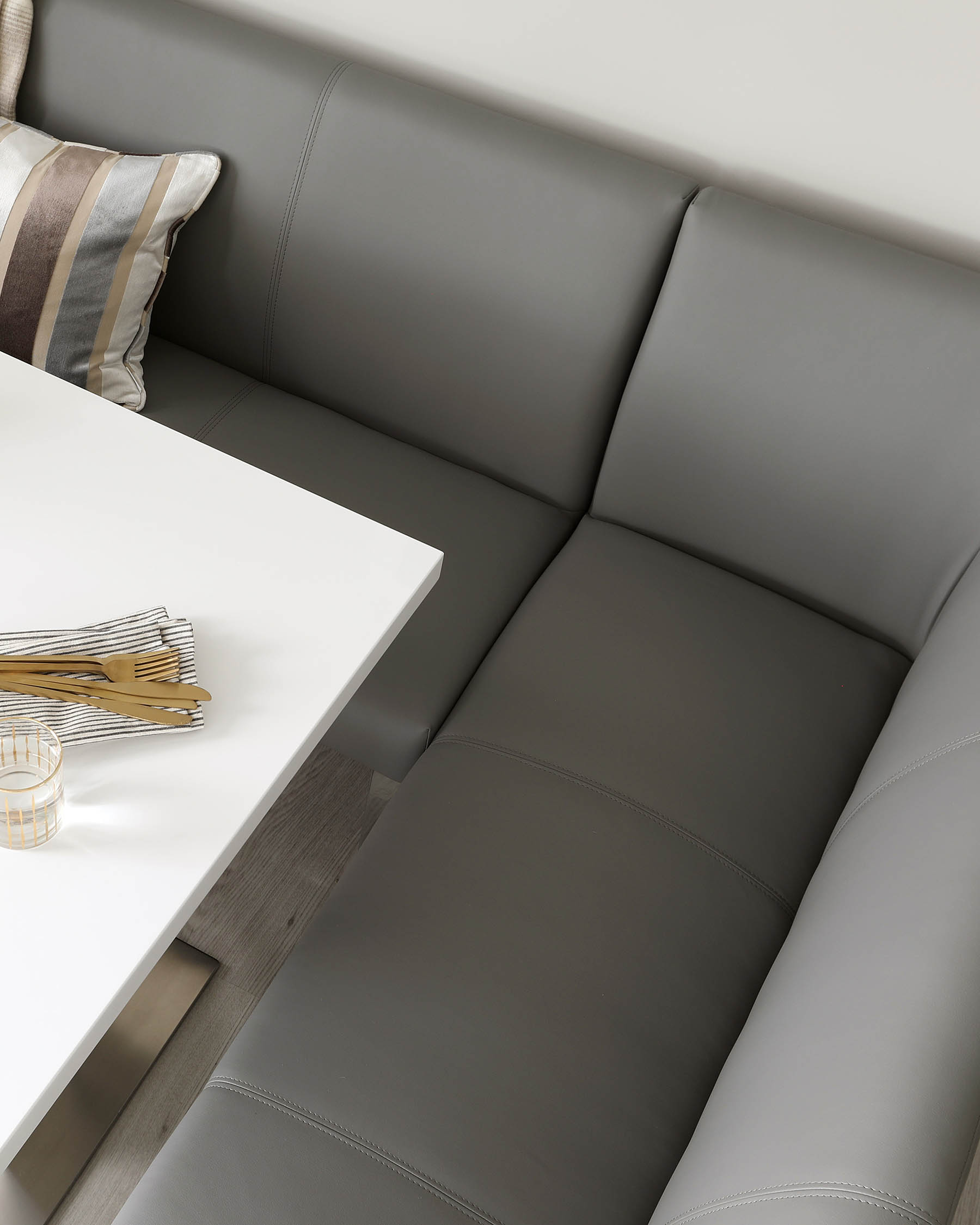 A modern corner bench with a seamless grey leather upholstery, featuring subtle stitch detailing and a plump cushioned seat, paired with a sleek white tabletop with a wooden base. A decorative striped pillow and a set of golden cutlery on a white napkin rest on the bench, beside a clear glass with gold embellishments.