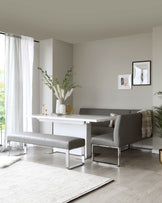 Modern minimalist dining room featuring a sleek white rectangular dining table, two grey upholstered benches with chrome legs, and a comfortable grey upholstered chair with chrome accents. A neutral-toned area rug lies beneath the arrangement on a light wooden floor, complementing the clean, contemporary aesthetic of the space.