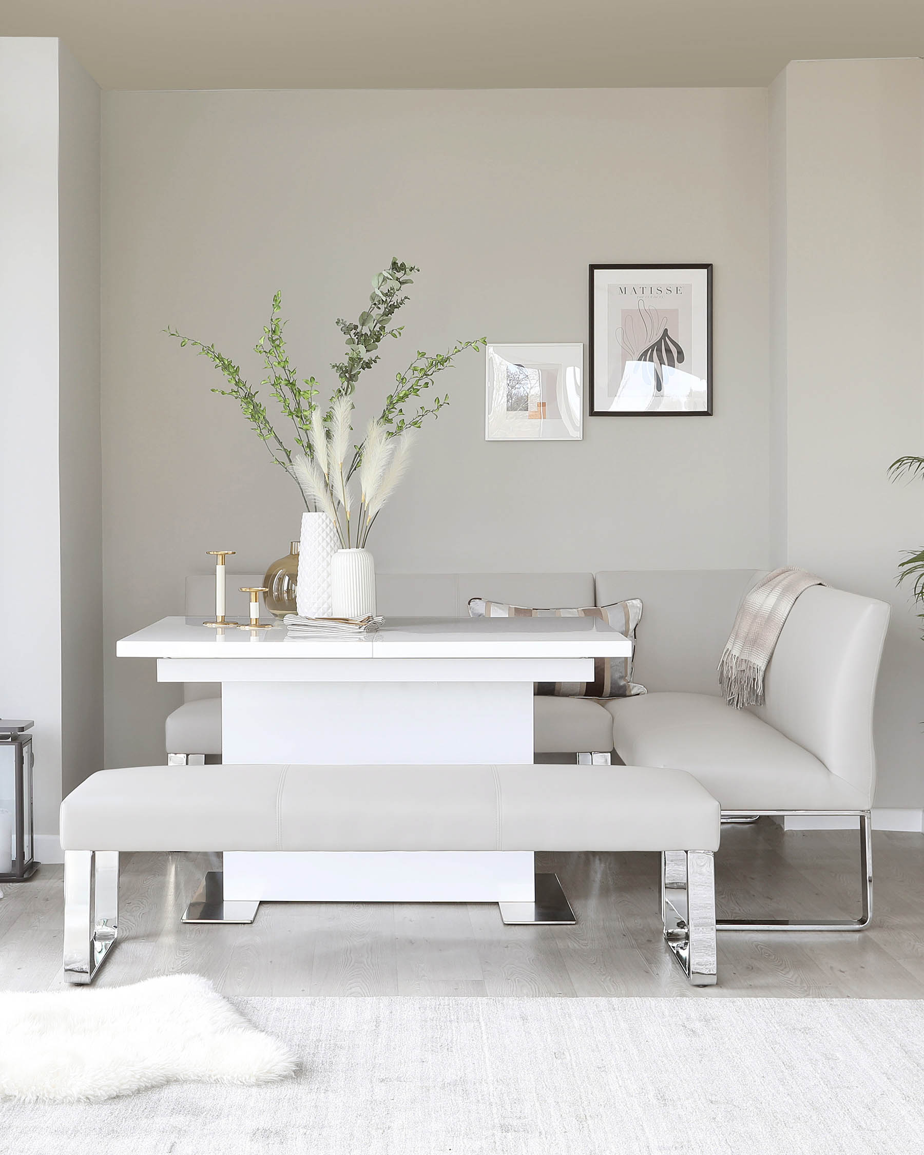 Modern white dining table with clean lines and glossy finish, paired with a matching white bench featuring chrome legs and a leather-like cushioned seat. The set is complemented by a leather-upholstered dining chair with a curved backrest and chrome cantilever base.