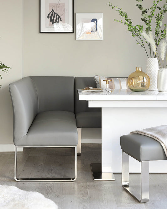 Modern dining nook with grey leather L-shaped bench and matching ottoman with chrome bases, paired with a white minimalist table.