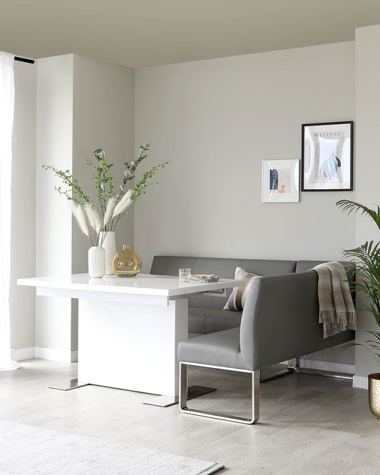 Modern minimalist dining set featuring a white rectangular table with a clean silhouette and metal base, paired with a sleek bench-style seat with grey upholstery and metal legs. A cosy throw blanket is draped over the bench, adding a touch of warmth to the scene.