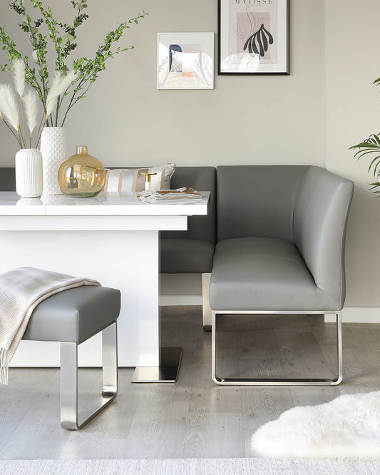 Modern minimalist dining area with a sleek white dining table paired with a grey upholstered bench and chair featuring clean lines and metallic bases. A cosy grey throw is draped over a simple stool, complementing the elegant colour palette of the room.