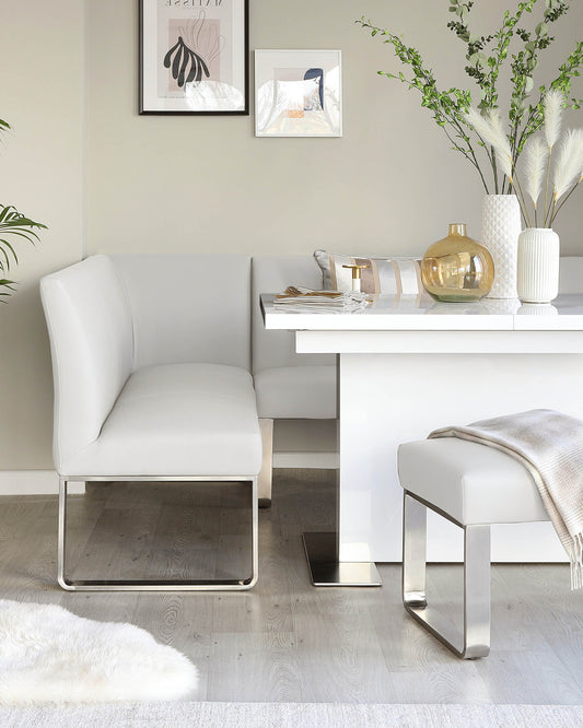 Modern minimalist furniture set with a white leather L-shaped dining bench featuring sleek chrome-plated legs, paired with a matching white leather stool. A white rectangular dining table holds decorative items, presenting a clean and contemporary aesthetic.