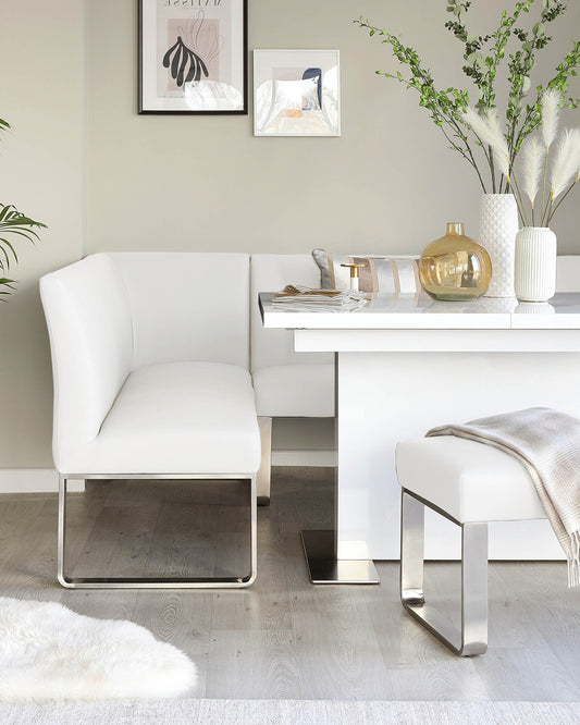 Modern minimalist dining set featuring a white corner bench with a sleek chrome base, paired with a matching white rectangular dining table also with a chrome foundation, complemented by a simple white dining stool with a chrome frame. The scene is accented by artistic wall decor and elegant vases with foliage for a sophisticated and clean interior aesthetic.