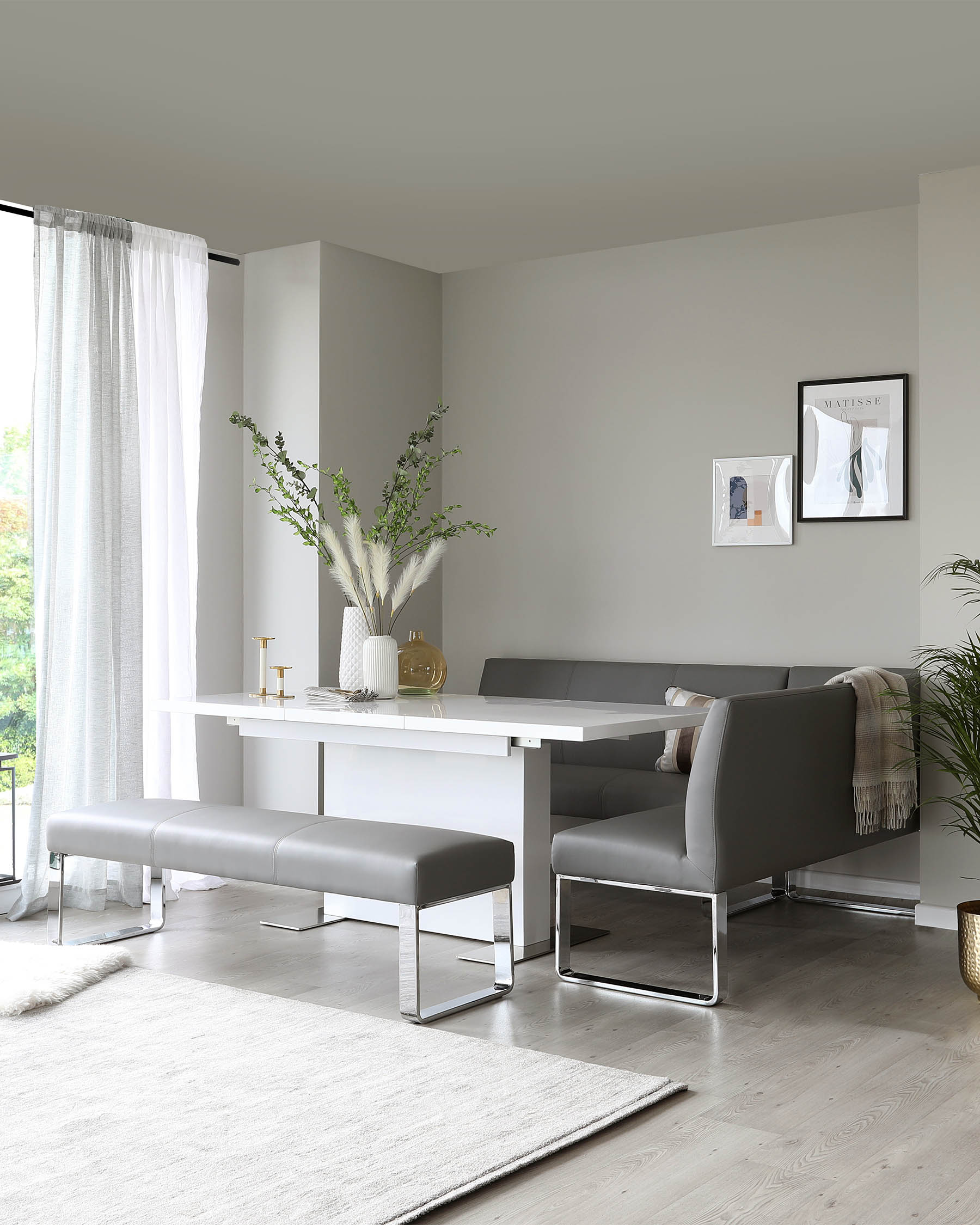 Modern minimalist dining set featuring a sleek white table with a wide, flat surface and square edges, accompanied by a dark grey upholstered bench and matching chairs with chrome legs. A plush off-white area rug lies beneath the set, enhancing the contemporary ambiance of the space.