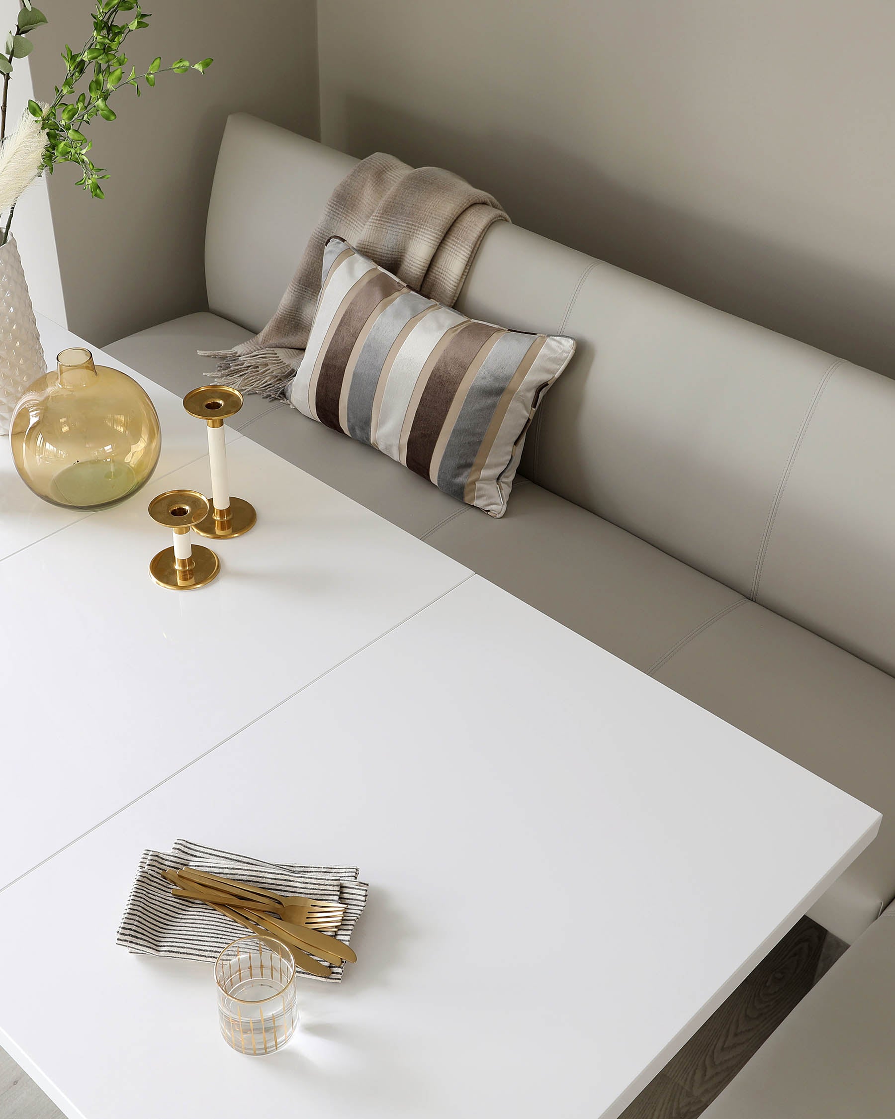 A modern, minimalist dining setup featuring a sleek white table with a glossy finish and a contemporary corner bench with light grey upholstery. The bench is accessorized with a striped cushion and a cosy beige throw.