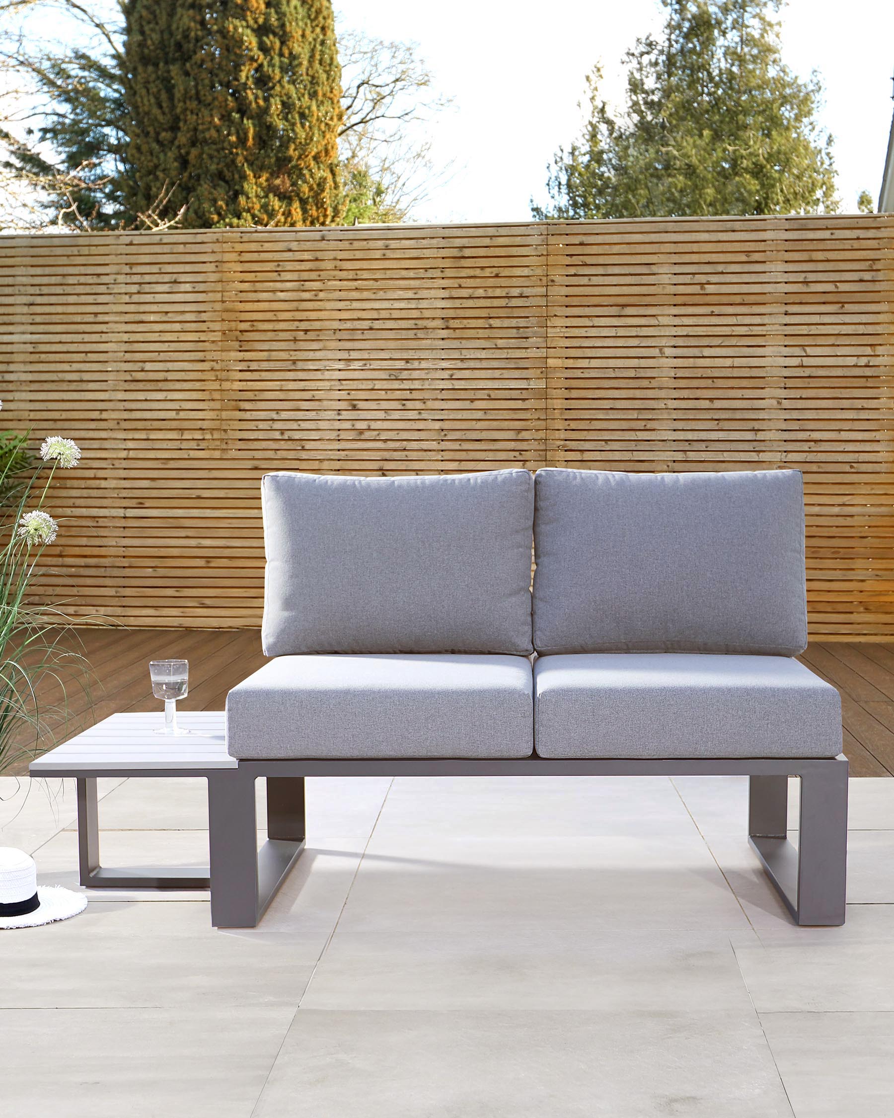 Modern outdoor furniture featuring a sleek two-seater sofa with grey upholstered cushions on a minimalist grey metal frame, paired with a small, square, white metal side table. The set is displayed on a patio with a wooden privacy fence in the background.