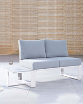 Modern two-seater outdoor sofa with minimalist white metal frame and light grey cushions, paired with a low-profile white metal frame coffee table with a clear glass top and a single glass resting on it, staged against a bamboo backdrop.