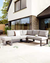 Modern outdoor sectional sofa with light grey cushions on a sleek dark metallic frame, accompanied by matching square ottoman and adorned with various neutral-toned throw pillows, positioned on a clean, paved patio area.