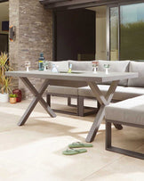 Modern outdoor dining area showcasing a minimalist rectangular table with a light grey top and distinctive X-shaped metallic legs, paired with a long, cushioned corner bench in a matching grey fabric.