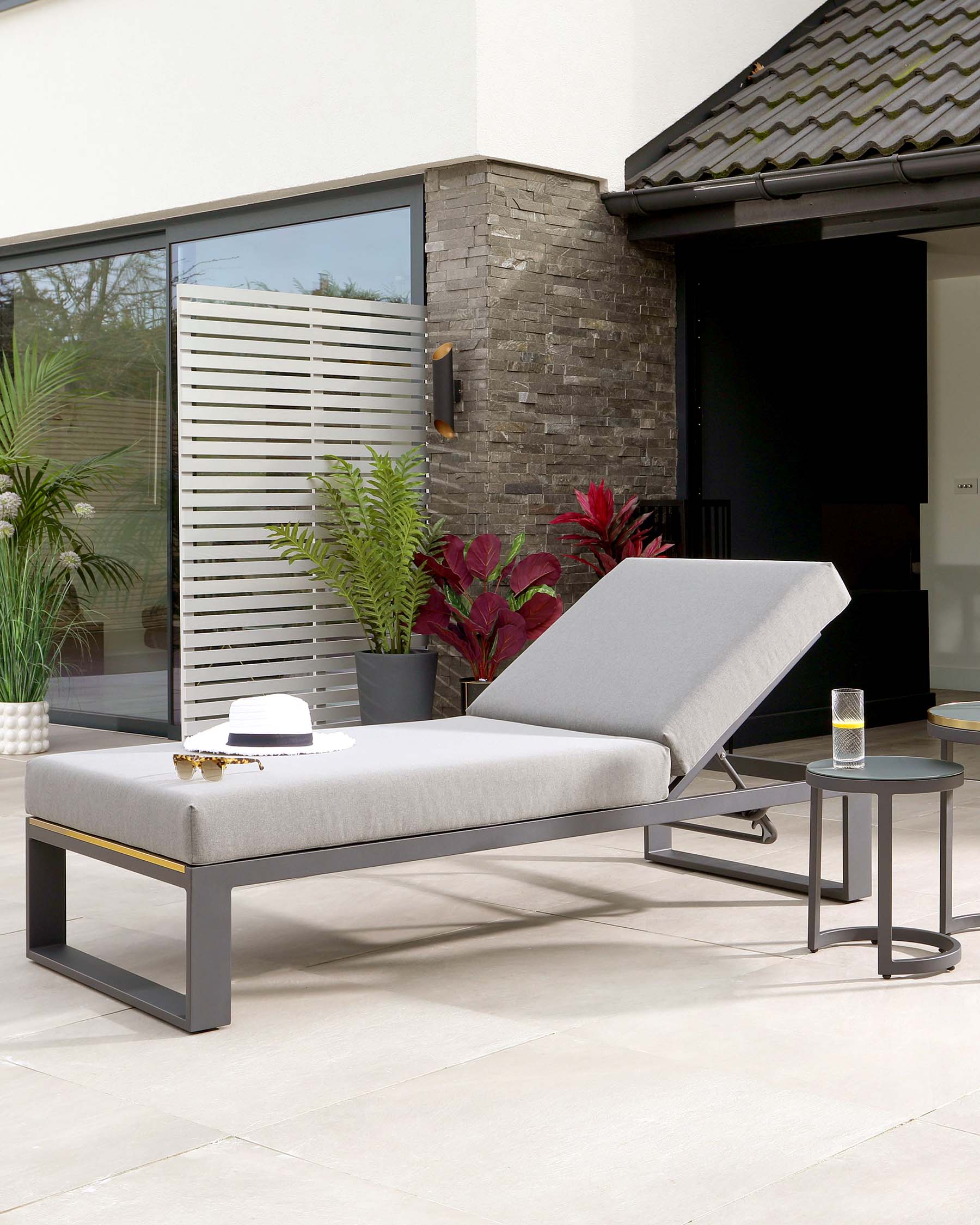 Modern outdoor patio furniture featuring a sleek chaise lounge with a light grey cushion on a dark metal frame, and a round side table with a matching dark metal frame and a dark tabletop.