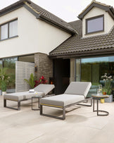 Modern outdoor furniture set featuring two minimalist-style, metal-framed loungers with light grey cushions, and a small, circular, metal side table with a dark glass top, displayed on a paved patio area.