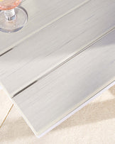 Close-up of a modern light grey wooden coffee table with a brushed finish on a beige carpet, featuring a section with fine wood grain details and a small part of a clear glass with a pink beverage on top.