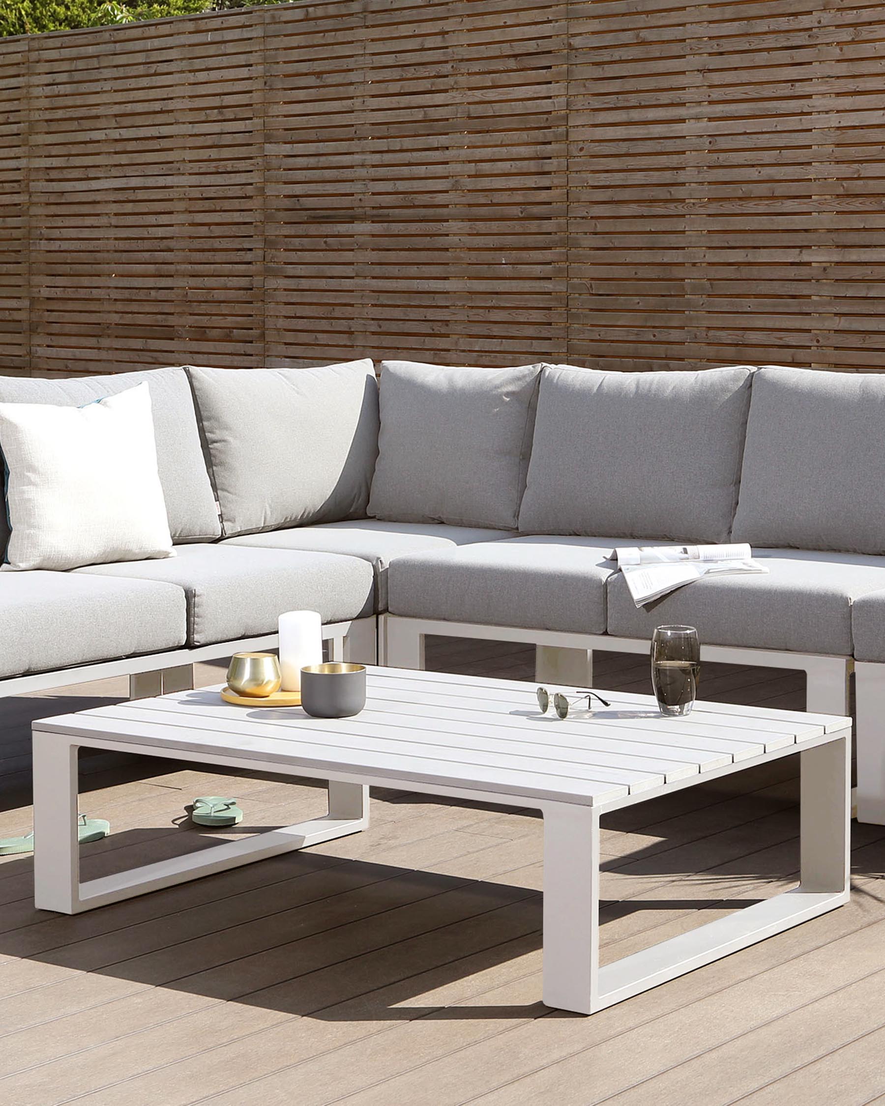 Outdoor patio furniture featuring a modern, white aluminium-framed sectional sofa with light grey cushions, complemented by a matching white rectangular coffee table with a slatted top and a lower shelf for additional storage. The set is positioned on a wooden deck with a bamboo privacy screen in the background, providing a contemporary and inviting outdoor lounge area.