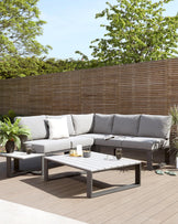 Modern outdoor furniture set featuring a sectional corner sofa with light grey cushions and a matching low-profile grey coffee table, displayed on a wooden deck and complemented by decorative green plants and accent pillows.