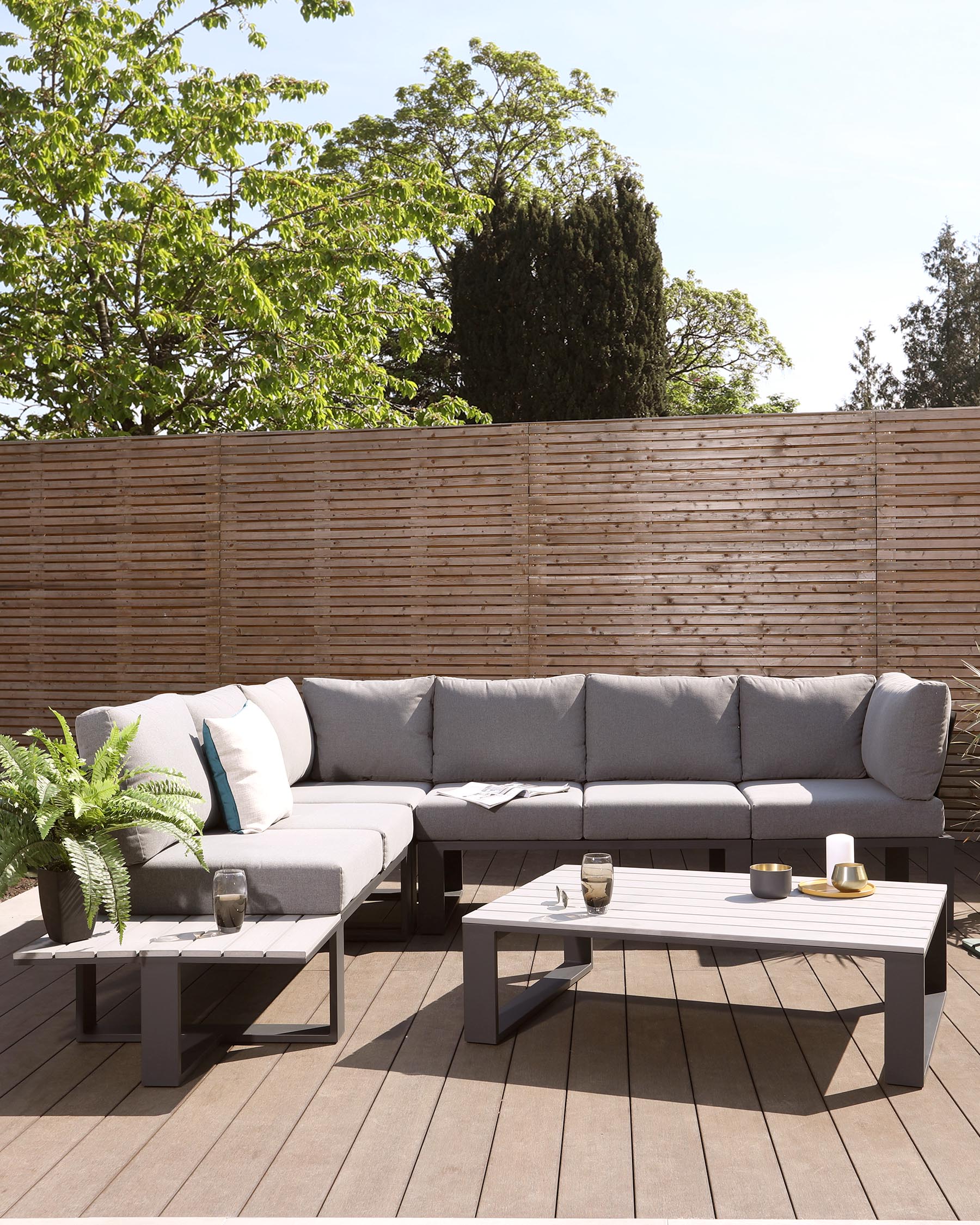 Modern outdoor L-shaped sectional sofa with light grey cushions and a sleek, low-profile, charcoal grey frame on a wooden deck. Accompanied by a matching rectangular coffee table with a slatted tabletop design. Decor includes assorted pillows, a fern plant, and candle holders on the table.