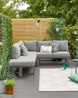 Outdoor garden furniture set featuring a modern design with a corner sectional sofa and a square coffee table, all with a dark grey metal frame. The cushions on the sofa are a light grey colour, adorned with decorative pillows in various patterns including stripes and abstract motifs. A geometric patterned outdoor rug in white and grey lies beneath the table. The setting is complemented with ambient string lighting and green plants.