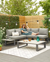 Modern outdoor furniture set featuring a grey sectional sofa with light grey cushions and an array of decorative pillows, complemented by a matching grey low-profile coffee table. Set is arranged on a textured outdoor rug with geometric pattern, showcasing a blend of comfort and contemporary design ideal for a patio or deck setting.