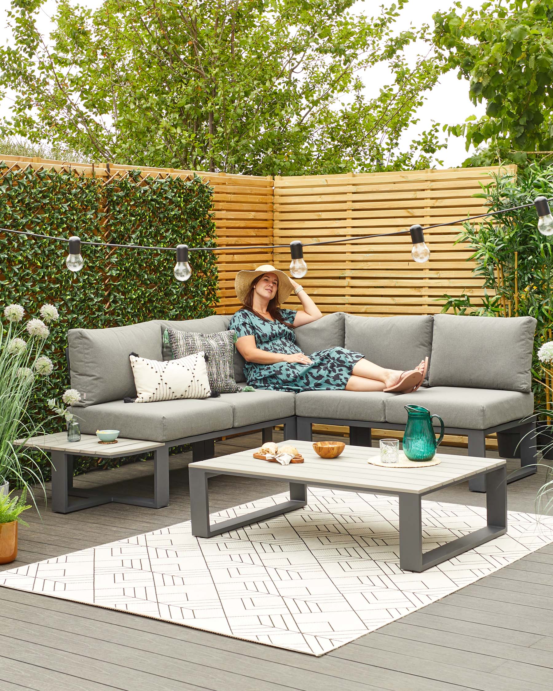 Modern outdoor furniture set featuring a grey sectional sofa with light grey cushions and an array of decorative pillows, complemented by a matching grey low-profile coffee table. Set is arranged on a textured outdoor rug with geometric pattern, showcasing a blend of comfort and contemporary design ideal for a patio or deck setting.