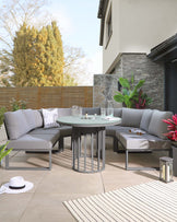 Modern outdoor living space with a corner sectional sofa in light grey fabric, accompanied by a round glass-top coffee table with a black metal base. A matching side table and decorative outdoor lanterns complement the setting, all arranged on a neutral, striped area rug.