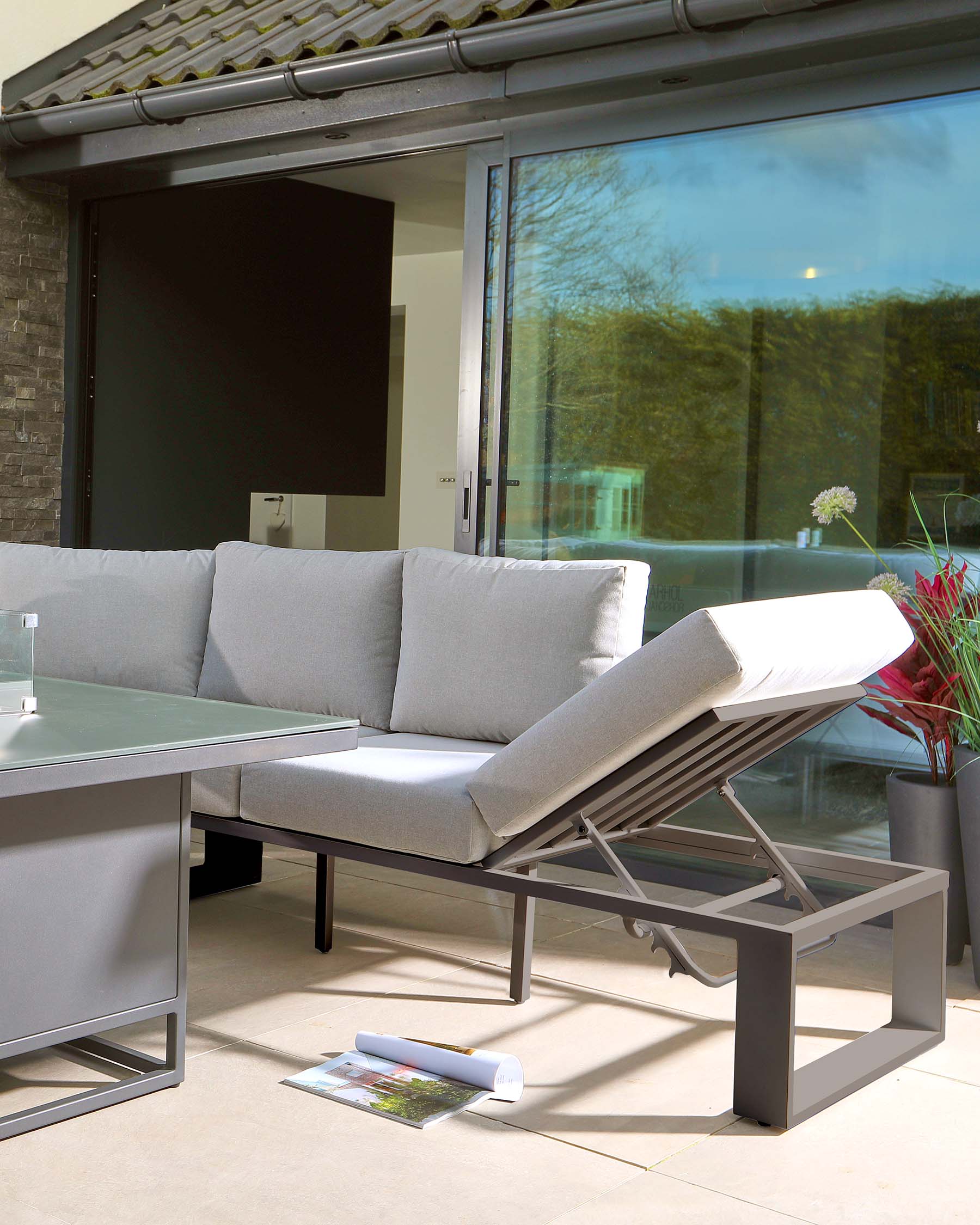 Modern outdoor furniture featuring a sleek, grey, aluminium frame sectional sofa with light grey cushions and a matching grey aluminium rectangular coffee table with a frosted glass top. The setting is complemented by a textured, light-tan outdoor area rug, with an open magazine resting casually on the ground, contributing to a relaxed, contemporary patio ambiance.