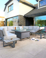 Modern outdoor patio furniture set with a grey sectional sofa featuring light grey cushions, a matching ottoman, and a square coffee table with a glass top, arranged on a paving stone floor.