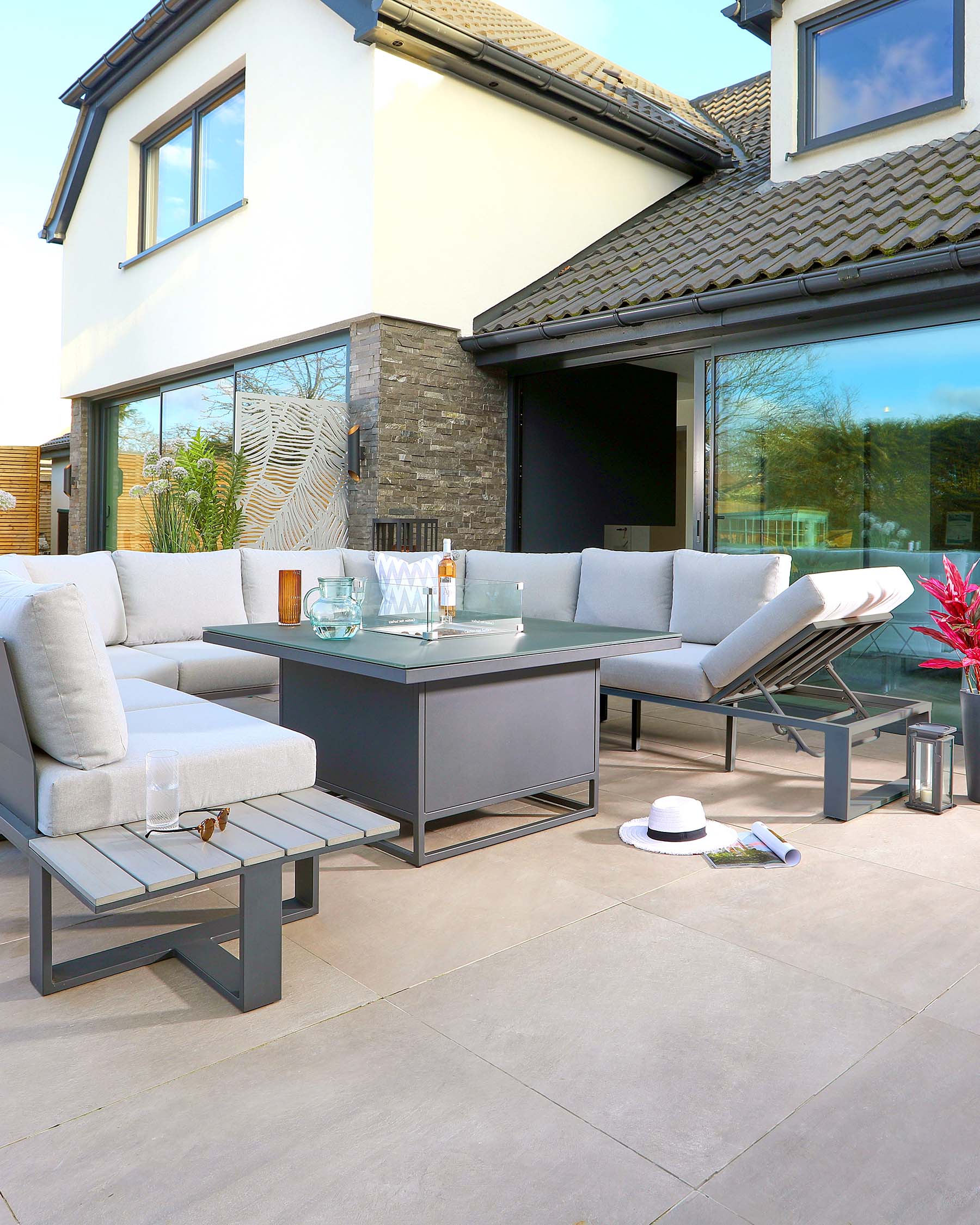 A modern outdoor lounge set featuring a sectional corner sofa with plush cushions, a rectangular centrepiece table with a glass top, and a matching square side table. The furniture is constructed with a sleek, grey metal frame, providing a contemporary and elegant look suitable for a patio setting.