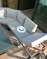 L-shaped outdoor sectional sofa with light grey cushions and a sleek metal frame, accompanied by a matching metal coffee table with a frosted glass top. A white hat and a magazine rest on the sofa.