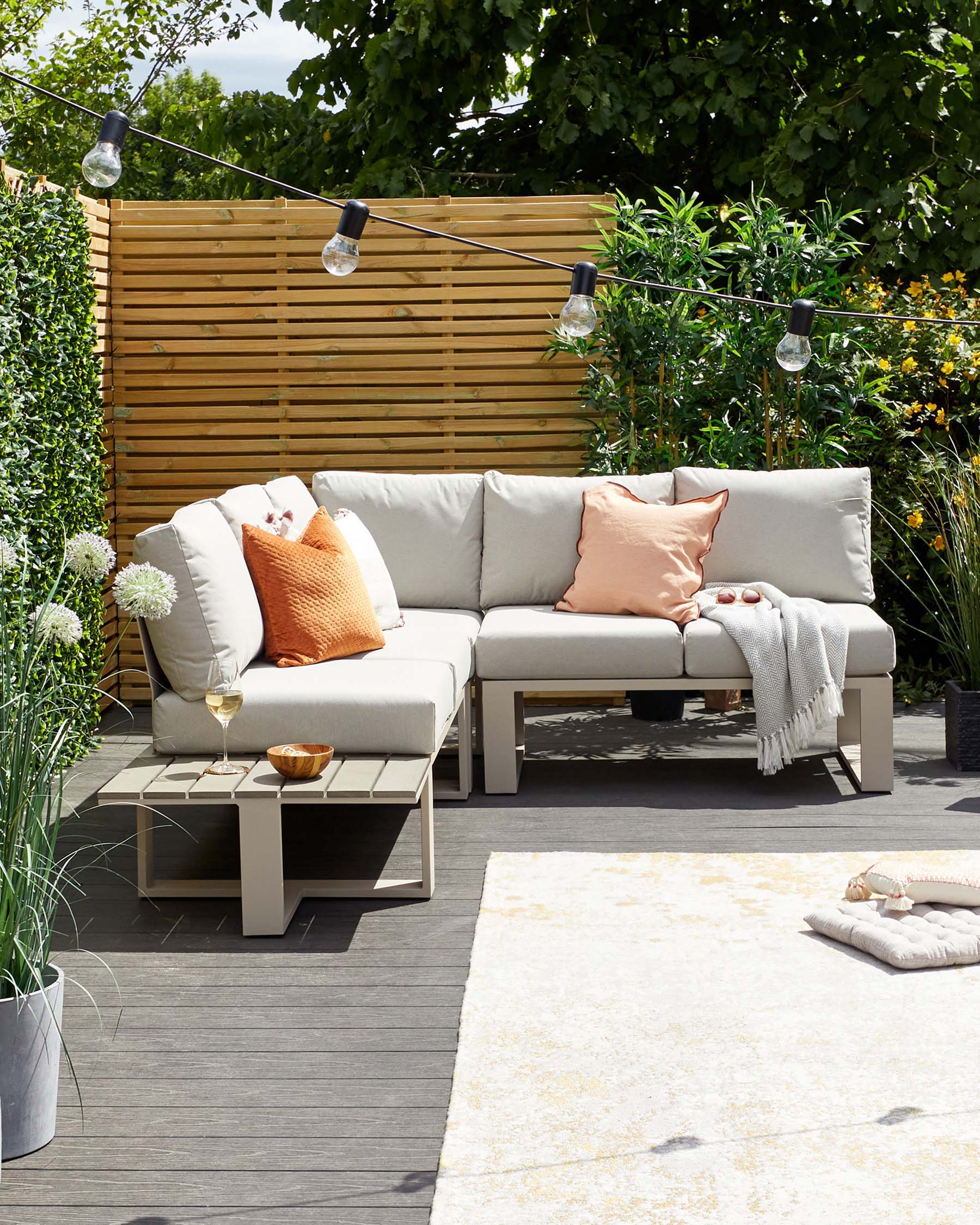 Modern outdoor garden furniture set featuring a beige modular sofa with cushions and a coordinating square coffee table, elegantly placed on a wooden deck with an accent rug. The setting is complemented by string lights overhead, adding a cosy ambiance.