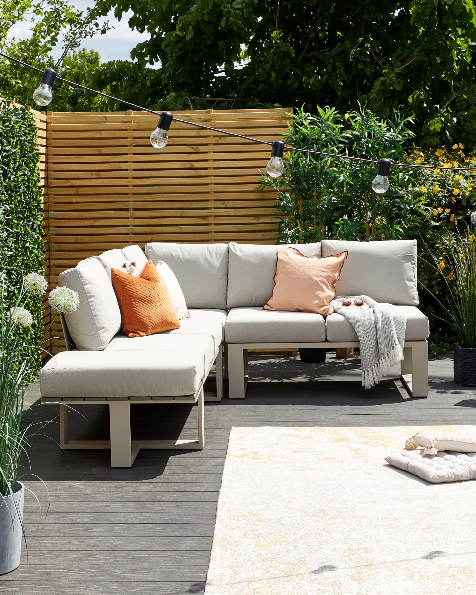 Outdoor furniture set with a modern design, featuring a sectional sofa with beige cushions and a matching ottoman, accompanied by a low coffee table. The set is placed on a grey decking area, complemented by decorative throw pillows and a light throw blanket. A cream and yellow area rug is partially visible in the foreground.