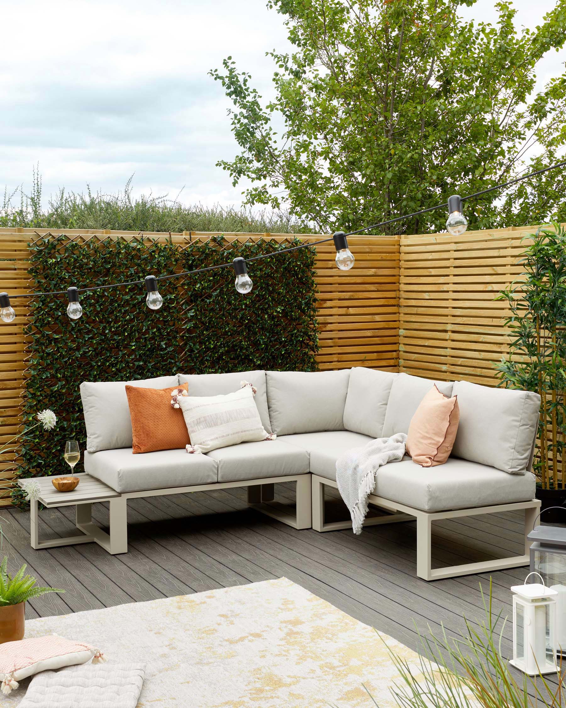 Modular outdoor sectional sofa in a light grey tone with white frames, adorned with pastel-coloured pillows, accompanied by a matching low side table, set on a wooden deck. A plush cream-and-gold area rug partially under the sofa and decorative lanterns complete the cosy setup.