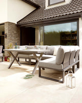 Outdoor furniture set featuring a modern grey sofa with clean lines and light grey cushions. Paired with the sofa is a rectangular white dining table with a unique X-shaped metallic base. The set presents a contemporary and elegant style perfect for patio or garden use.