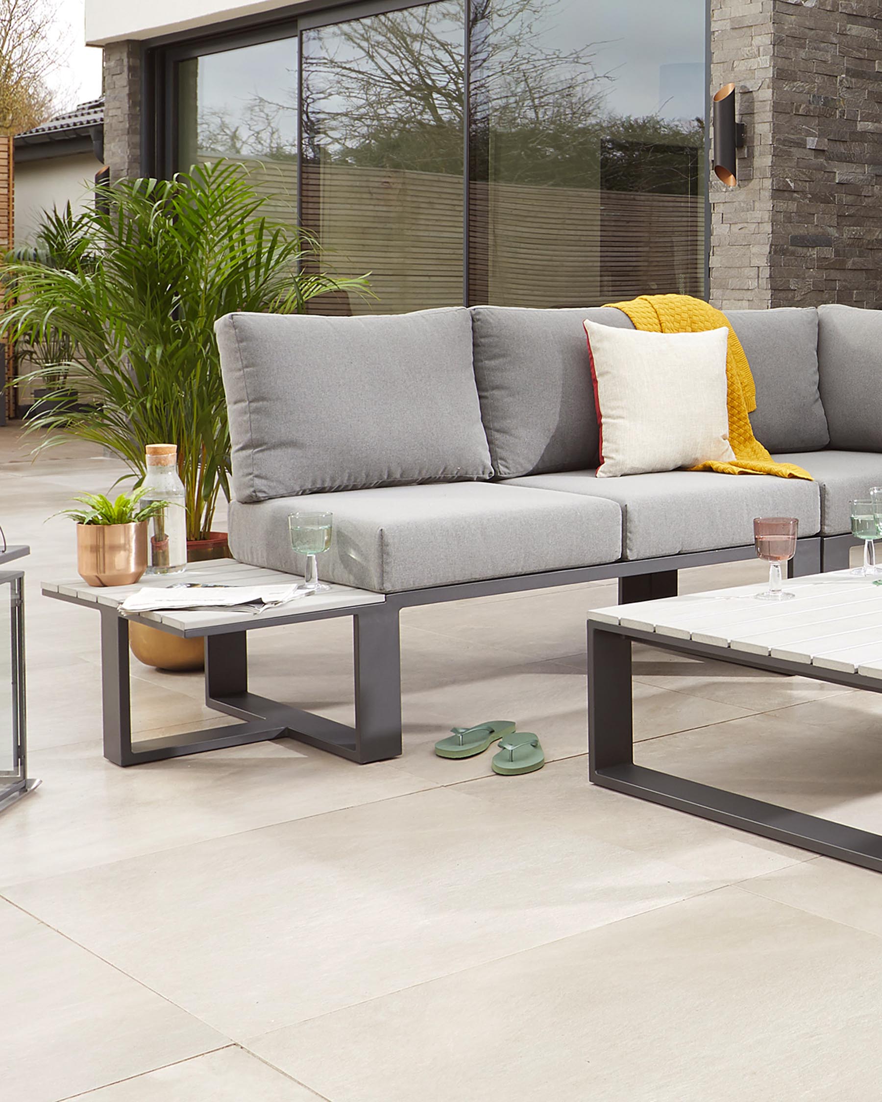 Modern outdoor furniture set featuring a sectional sofa with grey cushions and a low rectangular coffee table with a metal frame and white tile top. A side table with a round wooden top and metal frame is also visible. The set complements a contemporary patio setting.