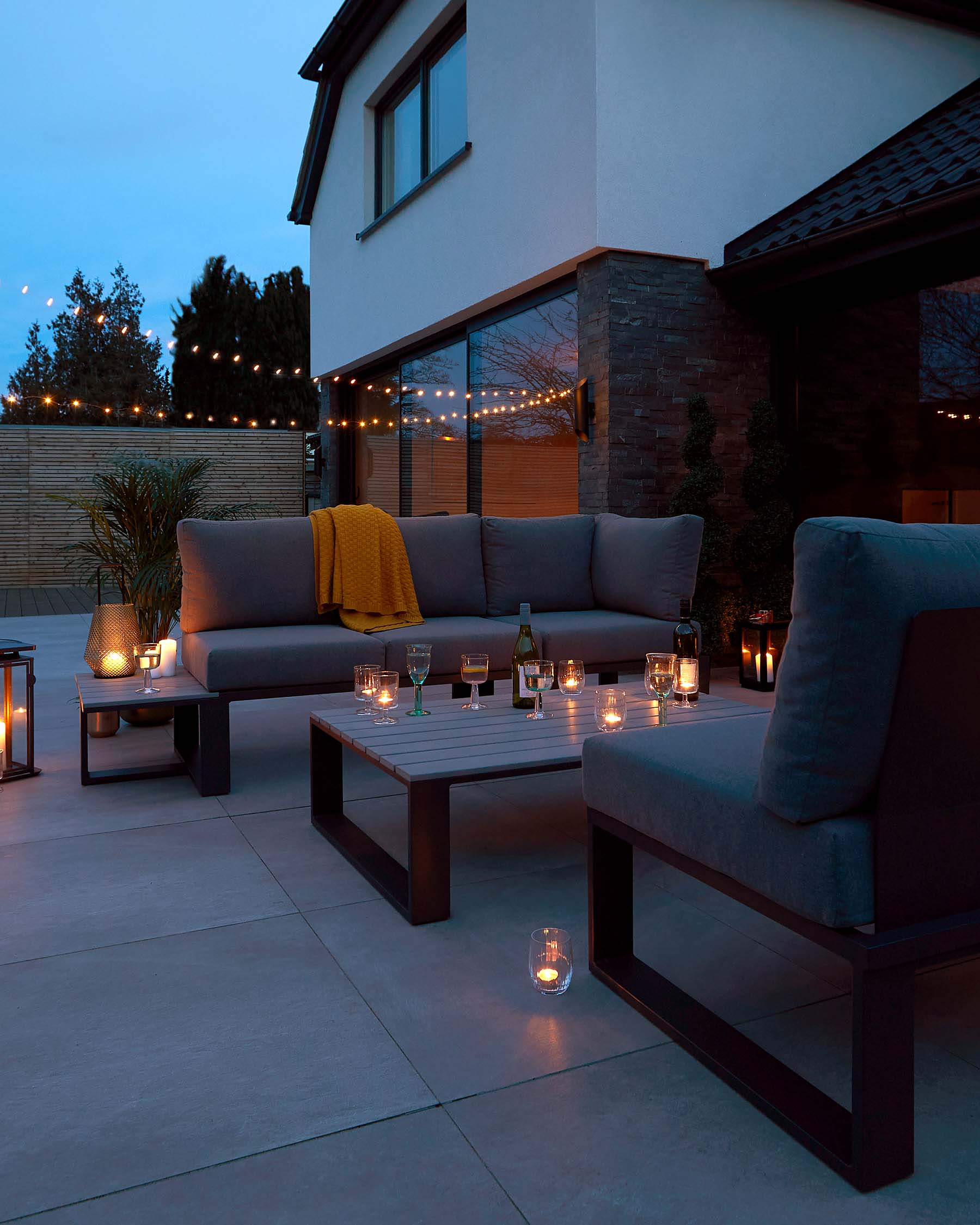 Outdoor patio set featuring a modern, sleek design with dark, wooden frames, including a three-seat sofa with comfortable grey cushions, a single armchair, and a rectangular coffee table. The furniture is complemented by a warm, yellow throw blanket, and the table is adorned with multiple lit candles inside glass holders, creating an inviting ambiance.