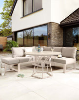 Modern outdoor furniture set featuring a round table with a geometric white base and a smooth tabletop, alongside an L-shaped sectional sofa with light taupe cushioning and white frames. The set includes matching throw pillows and is arranged on a patio.