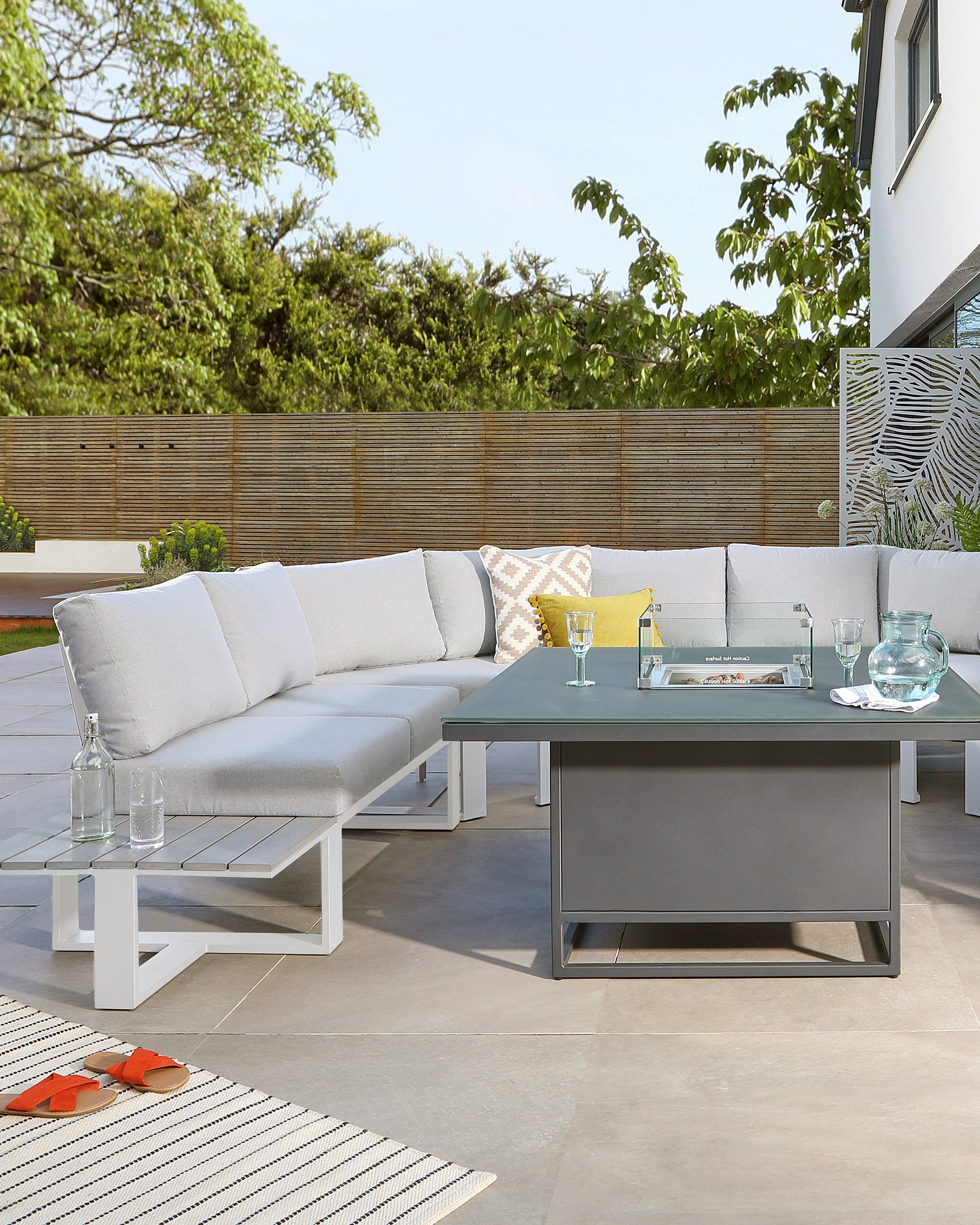 Modern outdoor furniture set featuring a sectional sofa with white aluminium frames and light grey cushions, accompanied by a rectangular metal coffee table with a dark finish. A striped outdoor rug lays beneath the setup, and decorative pillows and serving items adorn the table, creating a welcoming outdoor living space.