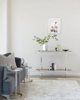 Modern minimalist living room with a pair of dark grey armchairs featuring plush cushions and silver metallic feet. Between the chairs are two small, round, glass-top side tables with silver bases. A sleek, rectangular glass-top console table with a metallic frame stands against the wall, adorned with decorative items such as books, a vase with greenery, and candles. A soft textured grey rug sits beneath the furniture ensemble.