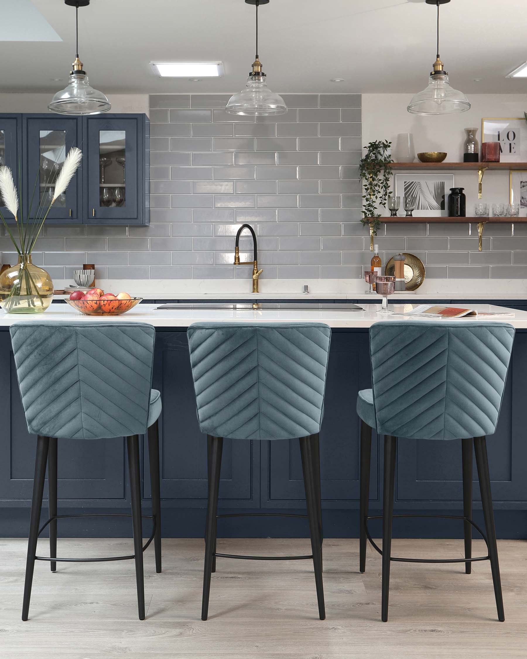 Three contemporary bar stools with plush teal velvet upholstery and chevron-style tufting detail. The stools feature a high-back design, slim black metal legs with footrest bars, and are positioned at a white countertop kitchen island.