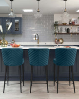 Three modern bar stools with teal, quilted velvet upholstery and black metal legs positioned at a kitchen island.